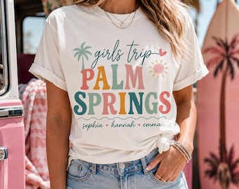 May include: A white short-sleeve t-shirt featuring the text girls trip PALM SPRINGS in colorful retro-style lettering. The design includes a palm tree, a sun, and a heart icon, with the names sophia, hannah, and emma printed at the bottom.