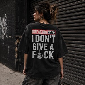 May include: Black, oversized t-shirt with the text "BREAKING NEWS I DON'T GIVE A F*CK" in white. The shirt has a distressed look and a crew neck. The text is in a bold, sans-serif font.