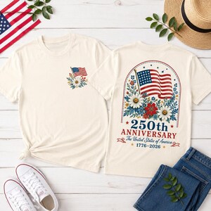 May include: Two cream-colored t-shirts with patriotic designs. One shirt features a small American flag with floral accents. The other displays a larger flag design with the text "250th Anniversary The United States of America 1776-2026".