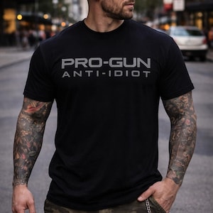 May include: Black t-shirt with the text "PRO-GUN ANTI-IDIOT" in gray. The person in the image is wearing a black cap, sunglasses, and camouflage cargo pants. The person has tattoos on their arms.