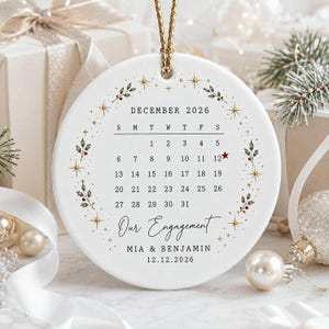 May include: A round white ceramic ornament featuring a December 2026 calendar with a red star on the 12th. The design includes a wreath of green holly leaves, red berries, and gold stars. Text reads Our Engagement, MIA & BENJAMIN, and 12.12.2026.