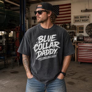 May include: A charcoal gray t-shirt with the text "BLUE COLLAR DADDY" in white brushstroke lettering. The shirt also features the text "DIRTY HANDS, BUSY NIGHTS". The person is wearing a black cap, sunglasses, and blue jeans.