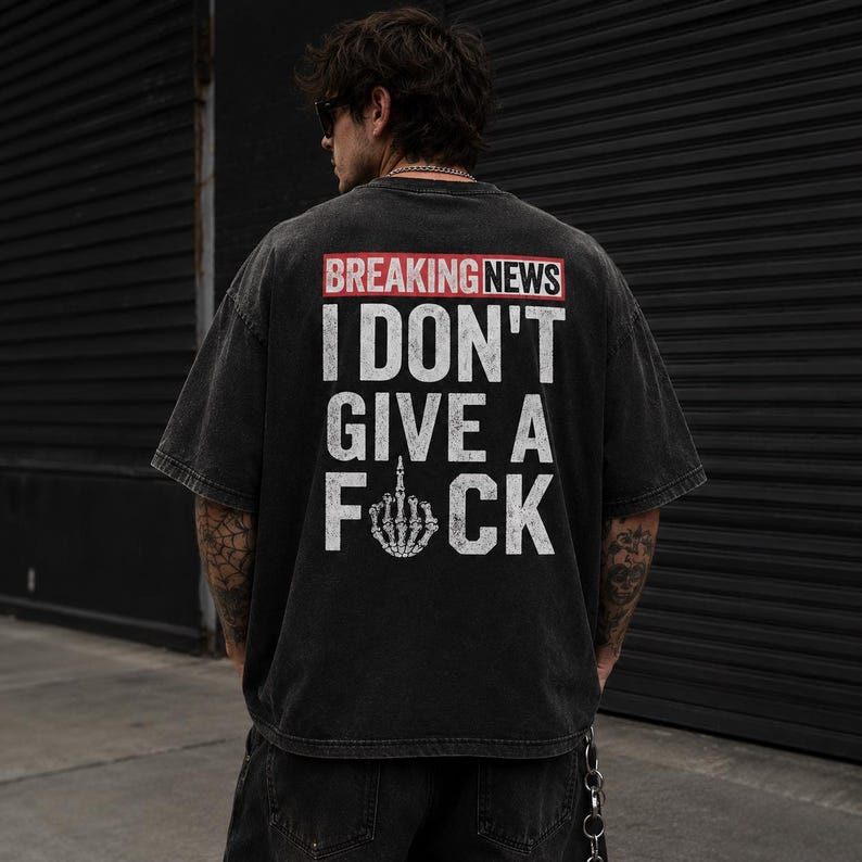 May include: Black oversized t-shirt with the text "BREAKING NEWS" in a red box, followed by "I DON'T GIVE A F*CK" in white block letters. The design includes a skeletal hand giving the middle finger.