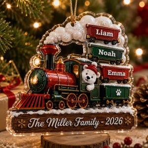 May include: A clear acrylic Christmas ornament shaped like a steam train with a white bear in the cabin. The train pulls cars labeled Liam, Noah, and Emma, with a paw print on the last car. A wooden base reads The Miller Family - 2026.