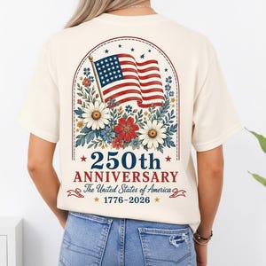 May include: Cream-colored t-shirt featuring a patriotic design for the 250th anniversary of the United States. The design includes the American flag, floral elements, and the text "250th Anniversary The United States of America 1776-2026".