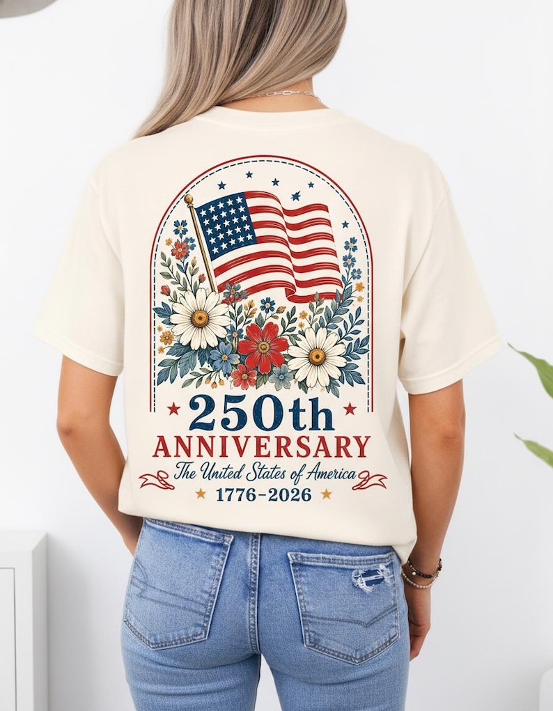 May include: Cream-colored t-shirt featuring a patriotic design for the 250th anniversary of the United States. The design includes the American flag, floral elements, and the text "250th Anniversary The United States of America 1776-2026".