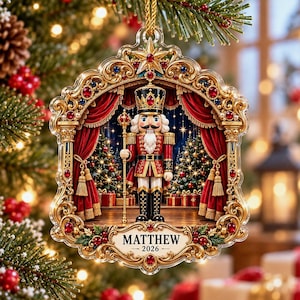 May include: A decorative Christmas ornament featuring a nutcracker soldier in a gold-trimmed frame. The nutcracker holds a staff, standing between red curtains. The base is inscribed with the name MATTHEW and the year 2026 in black text.