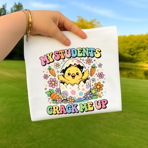 May include: White t-shirt featuring a colorful graphic of a baby chick hatching from an egg, surrounded by flowers and carrots. The text "MY STUDENTS CRACK ME UP" is displayed in a playful font. The design has a pastel color palette.