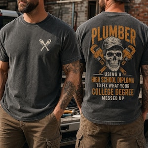May include: A charcoal gray short-sleeve t-shirt featuring a small graphic of two crossed pipe wrenches on the front chest. The back displays a large graphic of a skull wearing a cap, crossed wrenches, and the text PLUMBER USING A HIGH SCHOOL DIPLOMA TO FIX WHAT YOUR COLLEGE DEGREE MESSED UP.