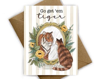 May include: Greeting card with a cat looking at its reflection in a mirror, which shows a tiger. The card has the text "Go get 'em tiger" in black script. The card is on a striped background with a brown envelope.