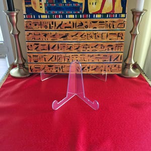 Puede incluir: Un caballete de acrílico transparente con tres patas, diseñado para exhibir una imagen o obra de arte enmarcada. El caballete está sobre una superficie roja.