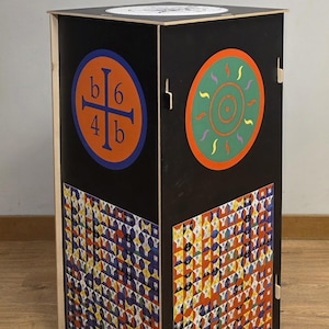 Op de afbeelding: Een geometrische bijzettafel met een zwarte buitenkant met kleurrijke cirkel- en kruisontwerpen. Het onderste deel van de tafel heeft een herhalend patroon van kleurrijke vormen. De tafel is gemaakt van hout en staat op een houten vloer.