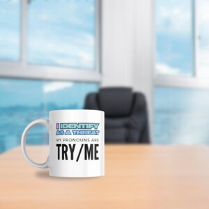 May include: White ceramic mug with the text "I IDENTIFY AS A THREAT, MY PRONOUNS ARE TRY/ME" in black, blue, and purple. The mug is on a light brown surface with a blurred office background.
