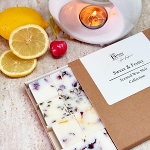 May include: A box of Sweet & Fruity scented wax melts with a white label. The wax melts are white with floral and herbal inclusions. A lit candle sits in a white ceramic oil warmer. Sliced lemons and a cherry are also present.