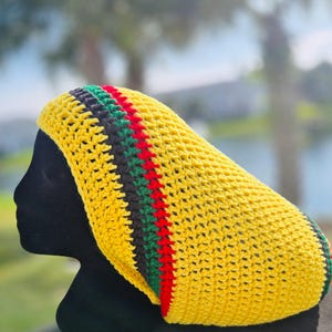 May include: A yellow crochet hat with a striped design in black, green, and red. The hat is displayed on a black mannequin head. The hat is a Rastafarian-inspired style.