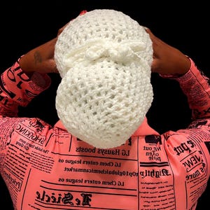 May include: A white crocheted hair snood featuring a textured, open-weave pattern and a decorative bow detail at the center. The accessory is worn over the hair, paired with a long-sleeved shirt printed with black newspaper-style text on a pink background.