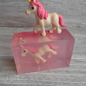 May include: A pink and white unicorn figurine with a yellow horn stands atop a pink, rectangular block. A similar unicorn is suspended within the block, creating a layered effect. The background is a light gray wooden surface.