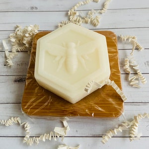 May include: A white hexagonal soap bar with a bee design on a wooden cutting board. The soap is wrapped in clear plastic.