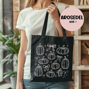 Peut inclure: Un sac fourre-tout noir orné d'un motif au trait blanc représentant diverses citrouilles, des nœuds et de petites étoiles. Le design est imprimé sur le devant du sac en tissu, doté de deux anses. Un logo rose circulaire indique AROSEDEL & CIE.