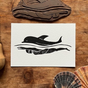 Könnte beinhalten: Ein Schwarz-Weiß-Linolschnitt eines Delfins, der im Meer schwimmt. Der Druck ist auf einer weißen Karte.