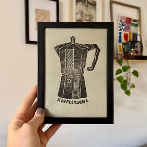 Könnte beinhalten: Schwarz gerahmter Druck mit einem Linolschnitt einer Espressokanne. Die Kaffeekanne ist dunkelbraun mit Griff und Ausguss. Der Text "KAFFEETSCH" ist am unteren Rand des Bildes aufgedruckt.