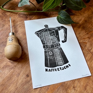 Könnte beinhalten: Ein schwarz-weiß Linolschnitt-Druck einer Espressokanne mit dem Wort "KAFFEETSCHI" darunter. Links vom Druck befindet sich ein Holzschnitzwerkzeug. Das Wort "COFFEE" ist unten links aufgedruckt.
