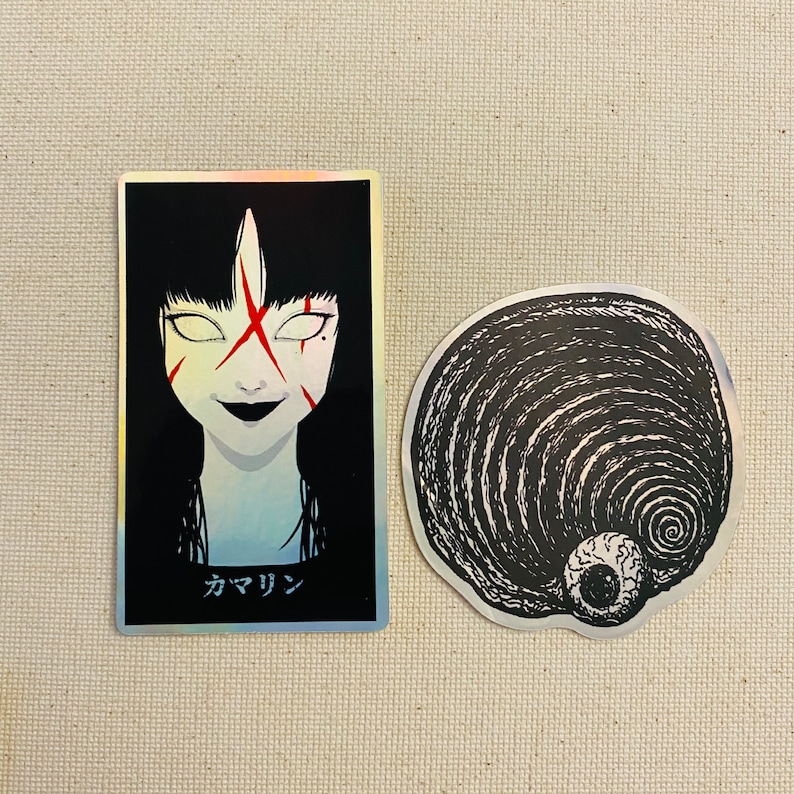 Może przedstawiać: Dwa naklejki, jedna przedstawiająca czarno-białą ilustrację kobiety z czerwonymi znakami na twarzy i tekstem "カマリン", druga przedstawiająca czarno-białą ilustrację spirali z okiem w środku.