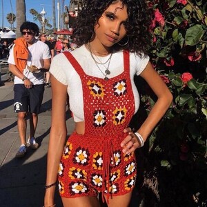 May include: A red crochet romper with daisy-like patterns in white, yellow, and brown. The romper features a high waist and suspender straps, paired with a white short-sleeved top. The background includes palm trees and a sunny outdoor setting.