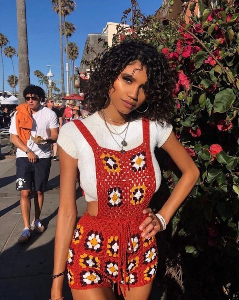 May include: A red crochet romper with daisy-like patterns in white, yellow, and brown. The romper features a high waist and suspender straps, paired with a white short-sleeved top. The background includes palm trees and a sunny outdoor setting.