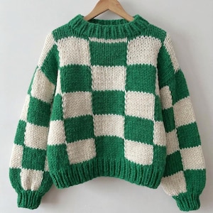 Op de afbeelding: Een grofgebreide trui met een groen en gebroken wit geblokt patroon, voorzien van een geribbelde ronde hals, boorden en zoom. De trui hangt aan een houten kledinghanger. De tekst onderaan luidt DOWNLOADABLE KNITTING PATTERN.