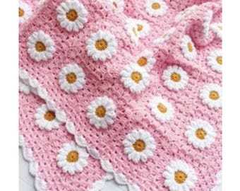 May include: A pink crocheted baby blanket with a white daisy pattern. The blanket has a scalloped edge.