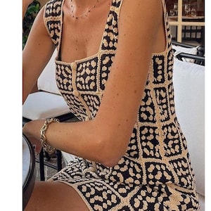 K&ouml;nnte beinhalten: Ein &auml;rmelloses Minikleid mit quadratischem Ausschnitt und einem sich wiederholenden Gittermuster. Das Kleidungsst&uuml;ck besteht aus geh&auml;kelten Quadraten in abwechselnden Farben Schwarz und Creme, mit einer zentralen vertikalen Knopfleiste.