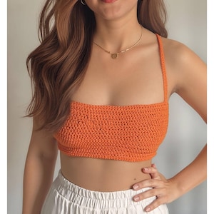May include: An orange crochet crop top with thin straps. The top is a solid colour and appears to be handmade. The model is wearing a gold necklace and white trousers. The top is a summer fashion item.