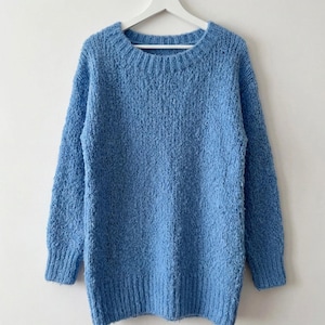May include: A bright blue long-sleeved sweater with a textured knit pattern, a ribbed crew neckline, and ribbed cuffs and hem. The sweater is displayed on a white hanger. The text at the bottom of the image reads DOWNLOADABLA KNITTING PATTERN.