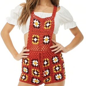 May include: A rust-colored crochet romper with a floral pattern. The romper features a drawstring waist and square neckline. The crochet design incorporates white, yellow, and brown flower motifs. The garment is worn over a white off-the-shoulder top.