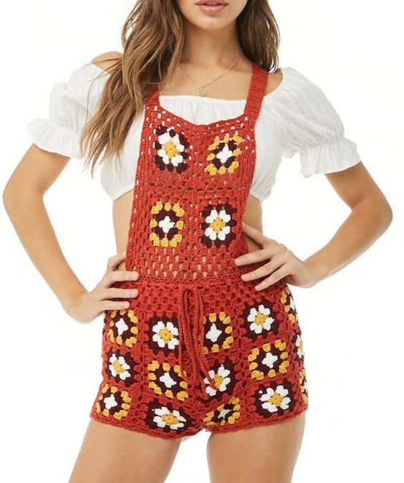 May include: A rust-colored crochet romper with a floral pattern. The romper features a drawstring waist and square neckline. The crochet design incorporates white, yellow, and brown flower motifs. The garment is worn over a white off-the-shoulder top.
