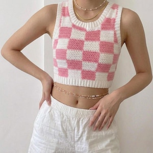 May include: A sleeveless cropped top featuring a pink and white chequered pattern made with crochet stitches. The garment has a round neckline and a ribbed hem. The text "DOWNLOADABLE CROCHET PATTERN" is printed in brown at the bottom of the image.