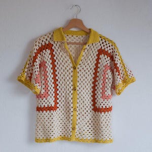 May include: Cream-colored crochet shirt with short sleeves and a yellow collar. The shirt features orange and pink geometric designs on the front. It has wooden buttons and is hanging on a wooden hanger.
