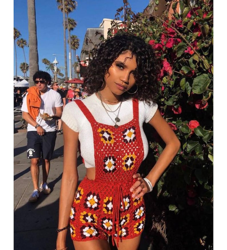 May include: A red crochet romper with daisy-like flower patterns in yellow, white, and black. The romper has a high waist and suspender straps. The model is wearing a white short-sleeved shirt underneath. The background shows palm trees and a sunny day.