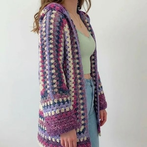 Può includere: Un cardigan con cappuccio all'uncinetto, con un motivo a quadrati granny. La giacca aperta presenta sfumature di viola, rosa e crema. Le maniche e il cappuccio sono di una tonalità viola più scura. Il cardigan è indossato sopra un top verde chiaro e jeans blu.