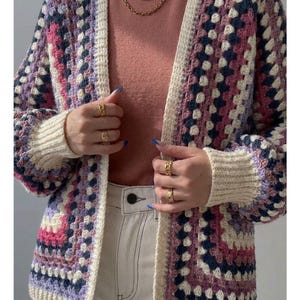 May include: A crochet cardigan featuring a granny square pattern in shades of purple, pink, navy blue, and cream. The garment has long sleeves with ribbed cream cuffs and a matching cream ribbed front border, worn over a solid pink shirt.
