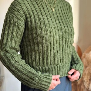 Könnte beinhalten: Ein langärmeliger olivgrüner Pullover mit vertikalem Rippstrickmuster, Rundhalsausschnitt und eng anliegendem Saum. Das Kleidungsstück ist in einer strukturierten Häkeloptik gefertigt und zeichnet sich durch eine einheitliche Farbe aus.