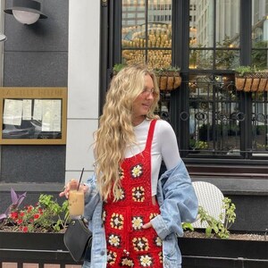 May include: Red crochet romper with white and yellow floral accents. The romper is worn over a white long-sleeve shirt and paired with a denim jacket. The outfit is accessorized with a black purse and a drink with ice.