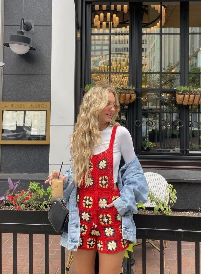 May include: Red crochet romper with white and yellow floral accents. The romper is worn over a white long-sleeve shirt and paired with a denim jacket. The outfit is accessorized with a black purse and a drink with ice.