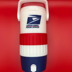 Può includere: Un refrigeratore per bevande in plastica bianca, rossa e blu con una maniglia bianca sulla parte superiore e un rubinetto bianco alla base. La parte anteriore mostra il logo blu dell'aquila dello United States Postal Service e il testo.