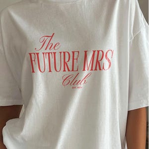 Puede incluir: Una camiseta blanca de manga corta con texto rojo en la parte delantera que dice The FUTURE MRS Club EST. 2025. La camiseta tiene cuello redondo y un corte holgado, con las palabras The y Club en letra cursiva y FUTURE MRS en letras de molde.