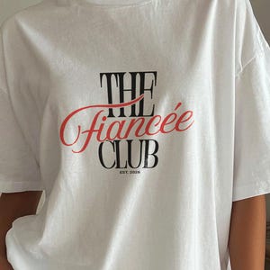 May include: A white short-sleeved t-shirt featuring a graphic print on the chest. The text reads THE in black, followed by the word fianc&eacute;e in a red cursive script, and the word CLUB in black, with the small text EST. 2026 printed below.