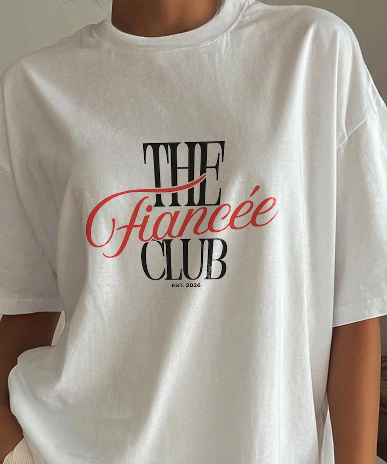 May include: A white short-sleeved t-shirt featuring a graphic print on the chest. The text reads THE in black, followed by the word fianc&eacute;e in a red cursive script, and the word CLUB in black, with the small text EST. 2026 printed below.