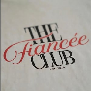 May include: A white t-shirt featuring black and red text that reads THE fianc&eacute;e CLUB EST. 2026. The word THE and CLUB are printed in a bold black serif font, while the word fianc&eacute;e is written in a red cursive script across the center.