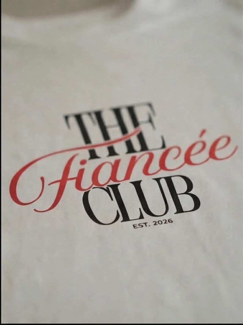 May include: A white t-shirt featuring black and red text that reads THE fianc&eacute;e CLUB EST. 2026. The word THE and CLUB are printed in a bold black serif font, while the word fianc&eacute;e is written in a red cursive script across the center.